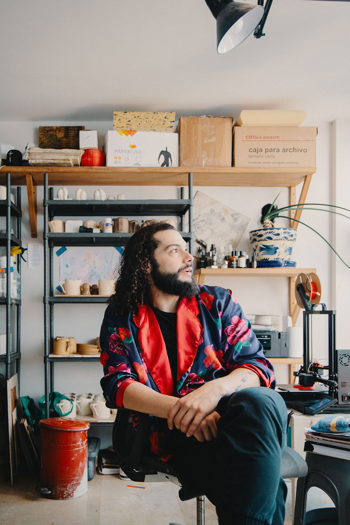 Man with dark curly hair and beard wearing floral jacket seated in a creative workspace with shelves.