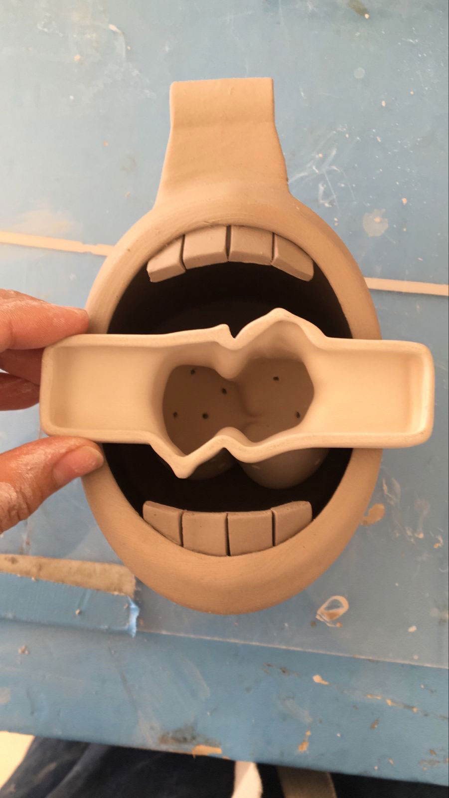Hand holding a ceramic mold above a round ceramic object shaped like an open mouth with teeth.