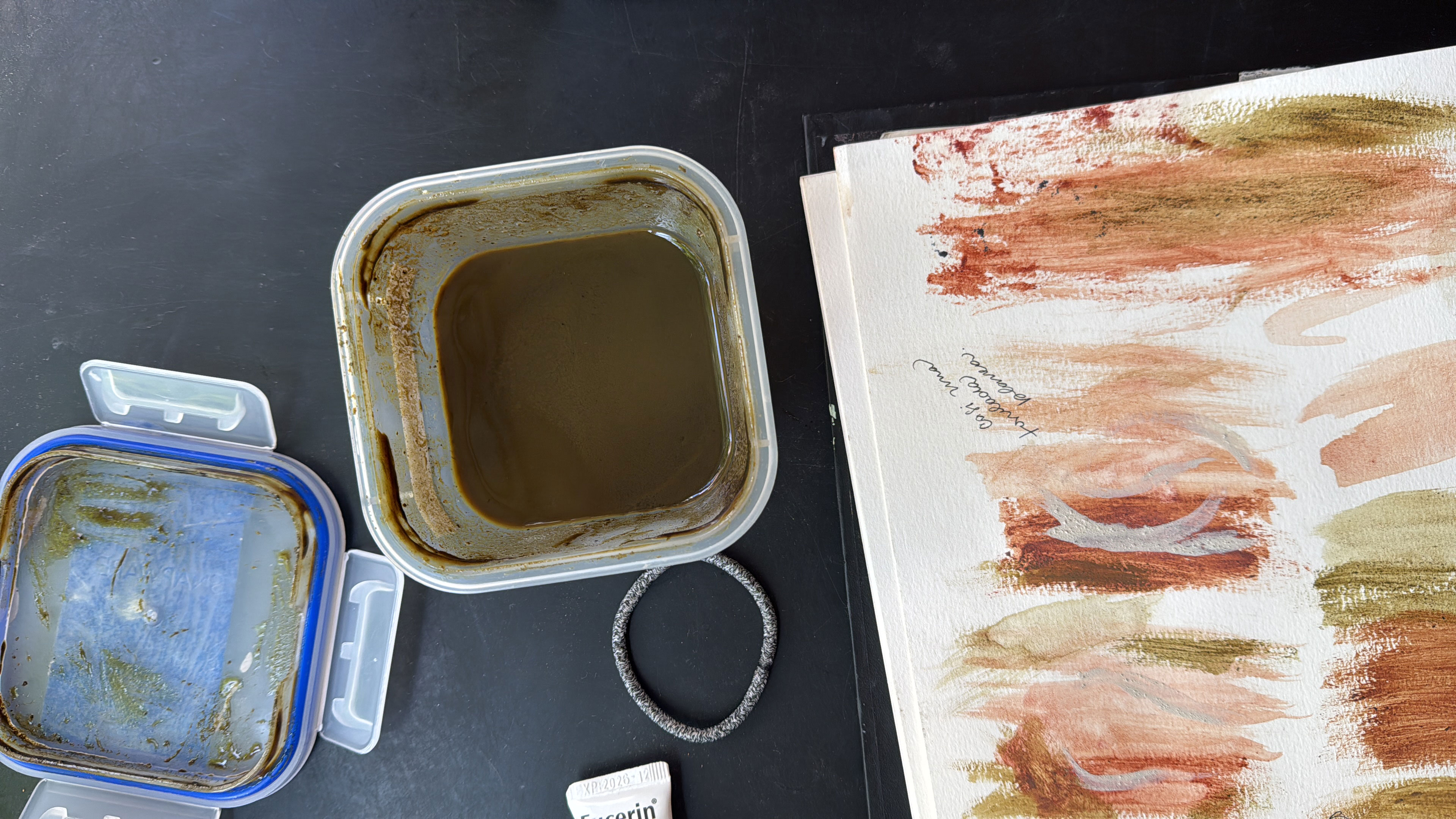 Open plastic container with brown liquid, hair tie, and watercolor paint swatches on paper.