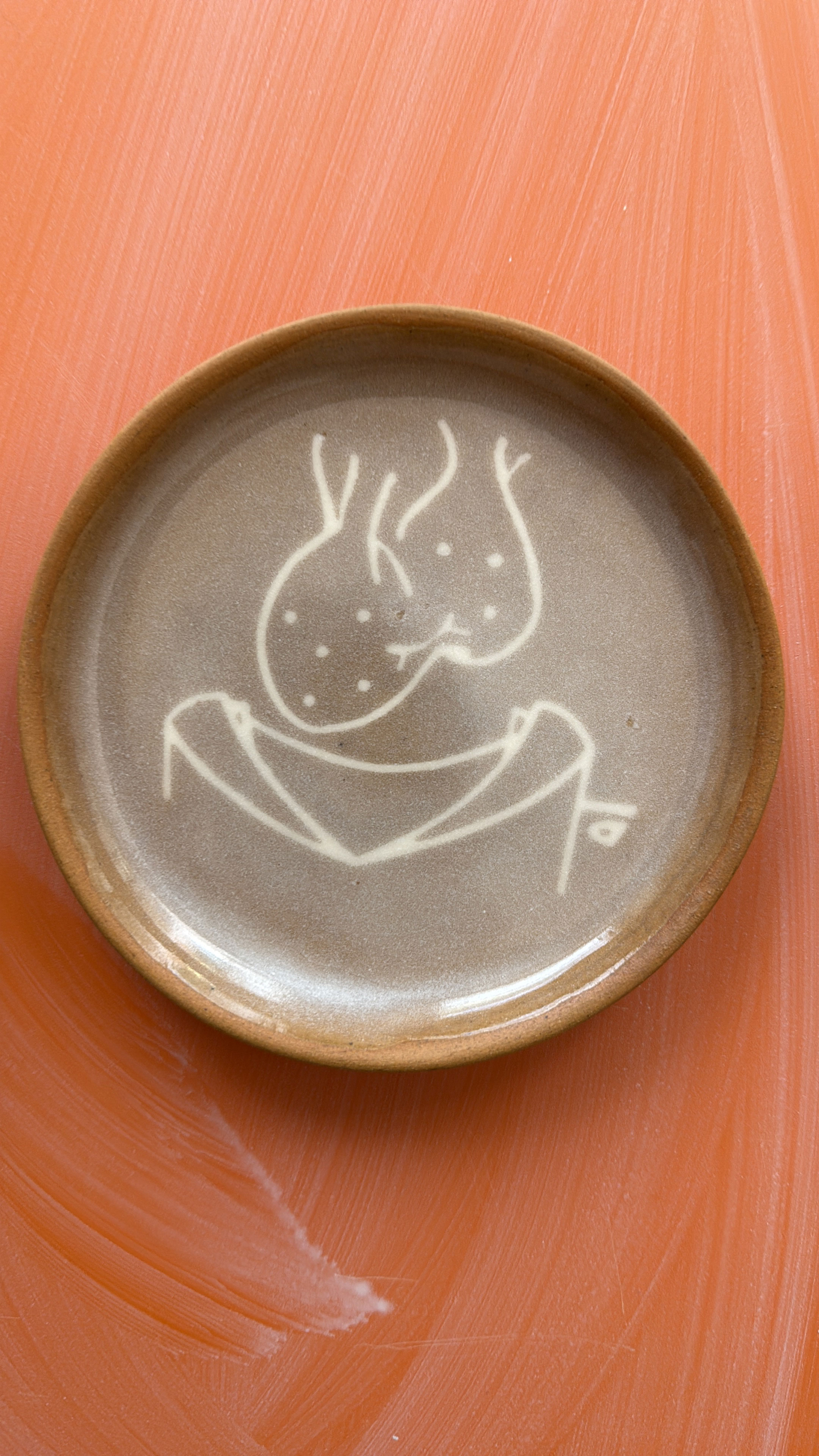Round ceramic plate with abstract white line art on a brownish-gray glaze, on an orange textured surface.