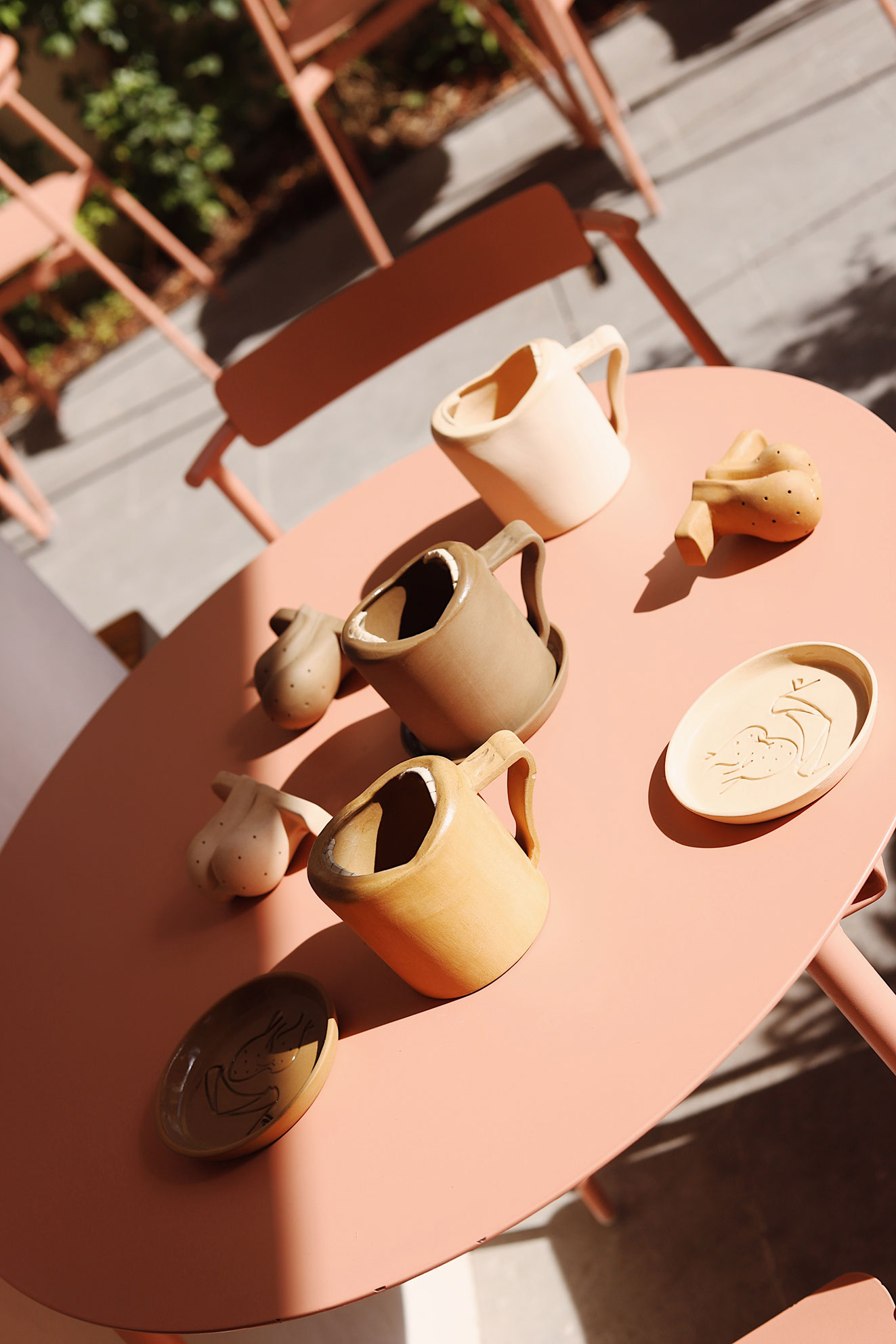 Three ceramic mugs, two plates, and three small ceramic figures on a round salmon-colored table outdoors.