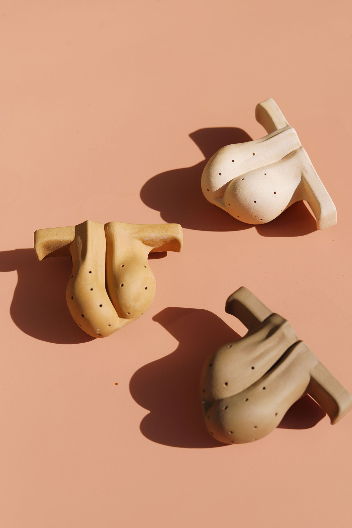Three ceramic sculptures of male genitalia in different earth tones on a peach background.