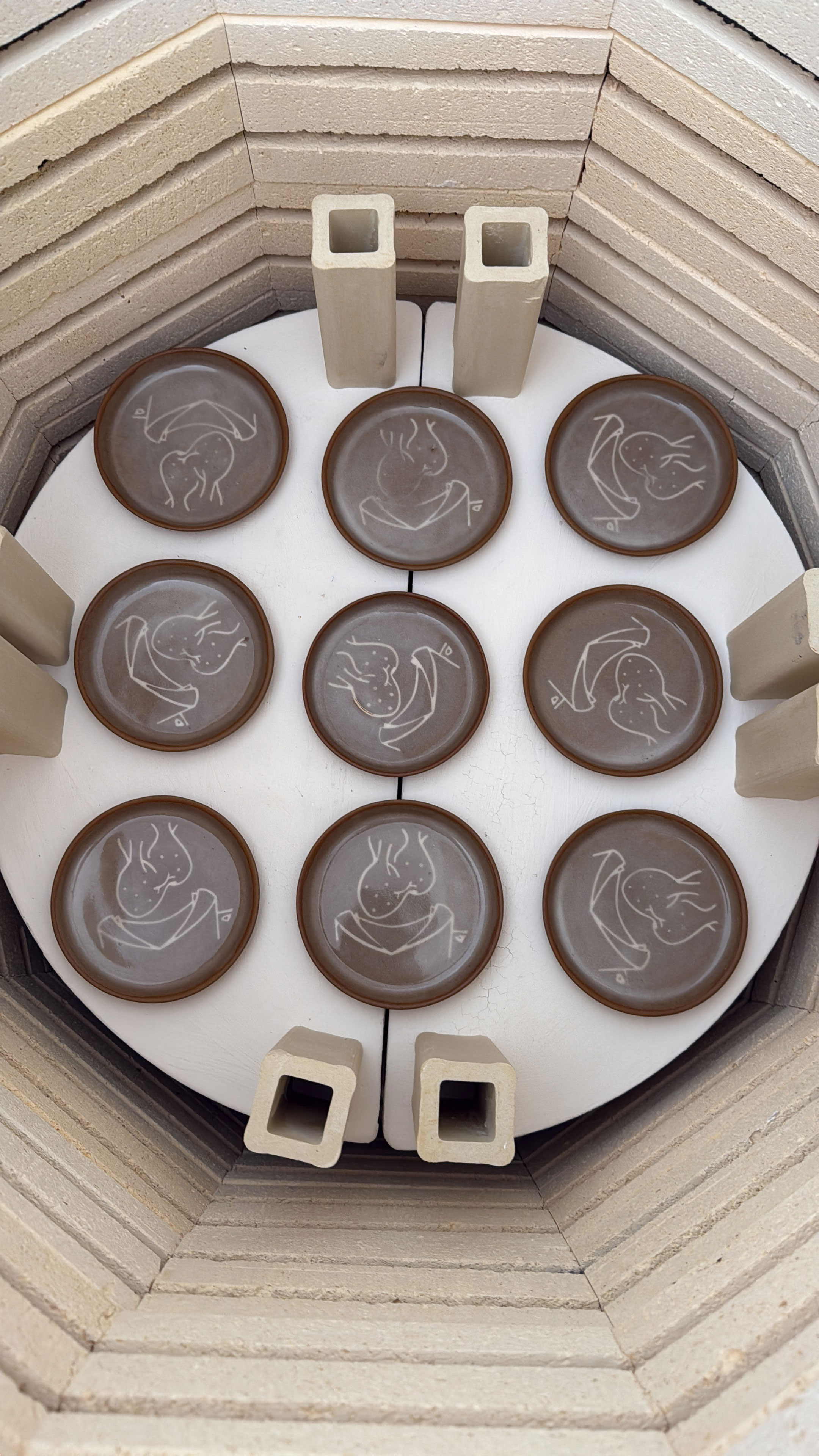 Nine brown ceramic plates with white abstract designs arranged in a kiln with four beige ceramic posts.