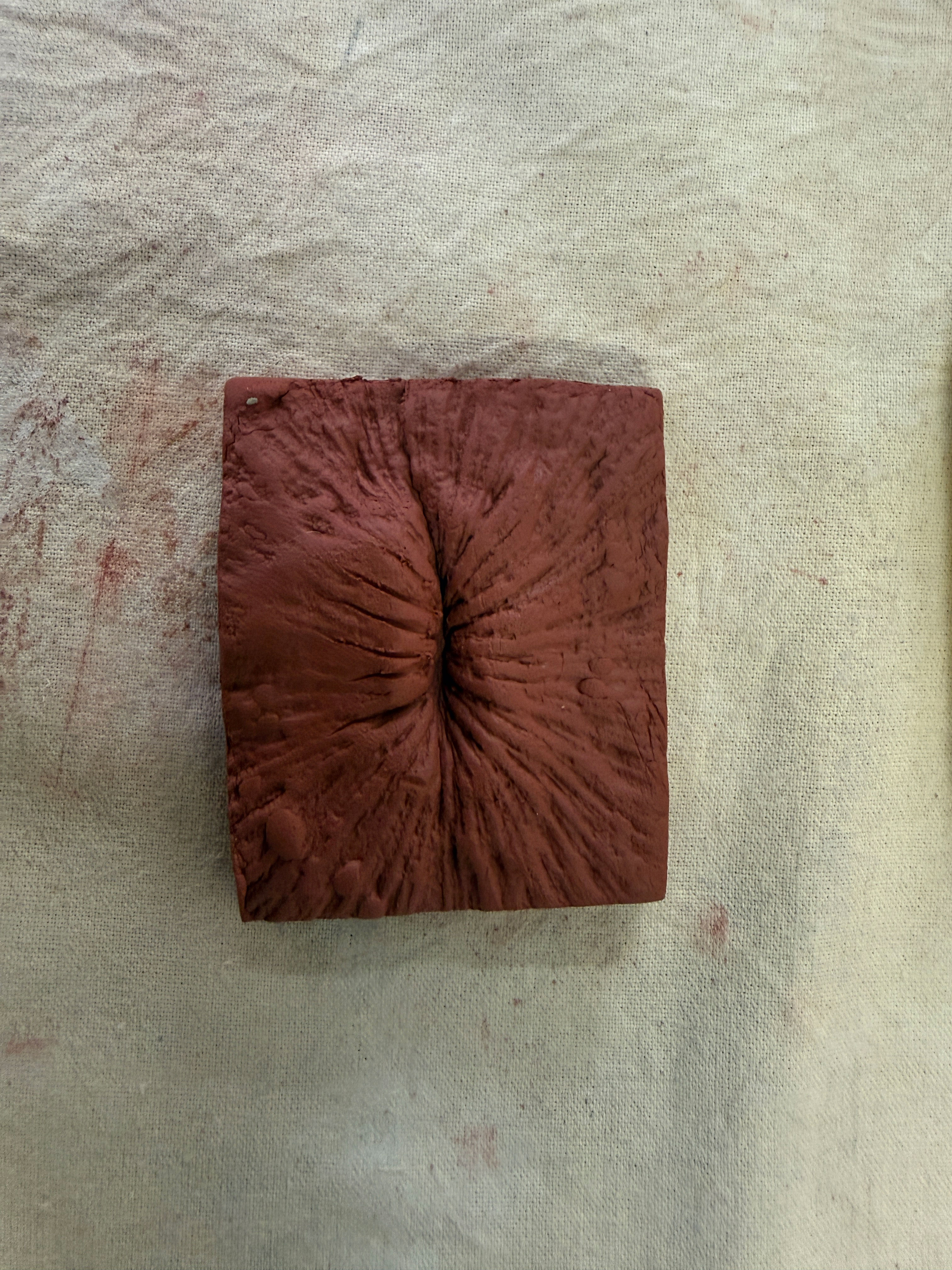 Square-shaped textured brown clay piece with deep central indentation and radiating lines.