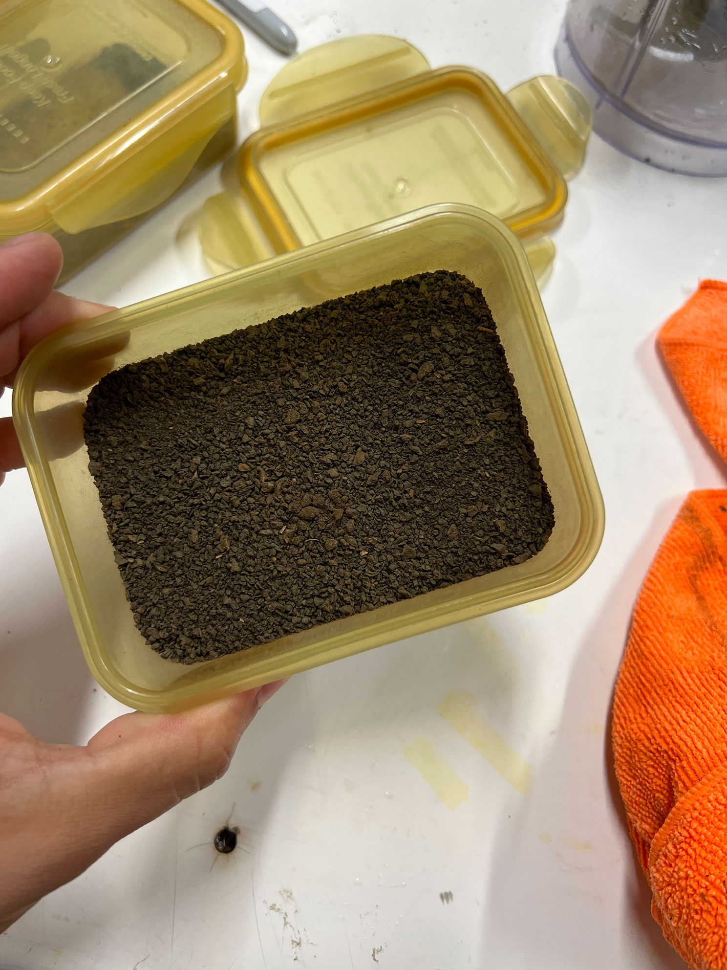 Hand holding a yellow container filled with dark brown coffee grounds on a white surface.