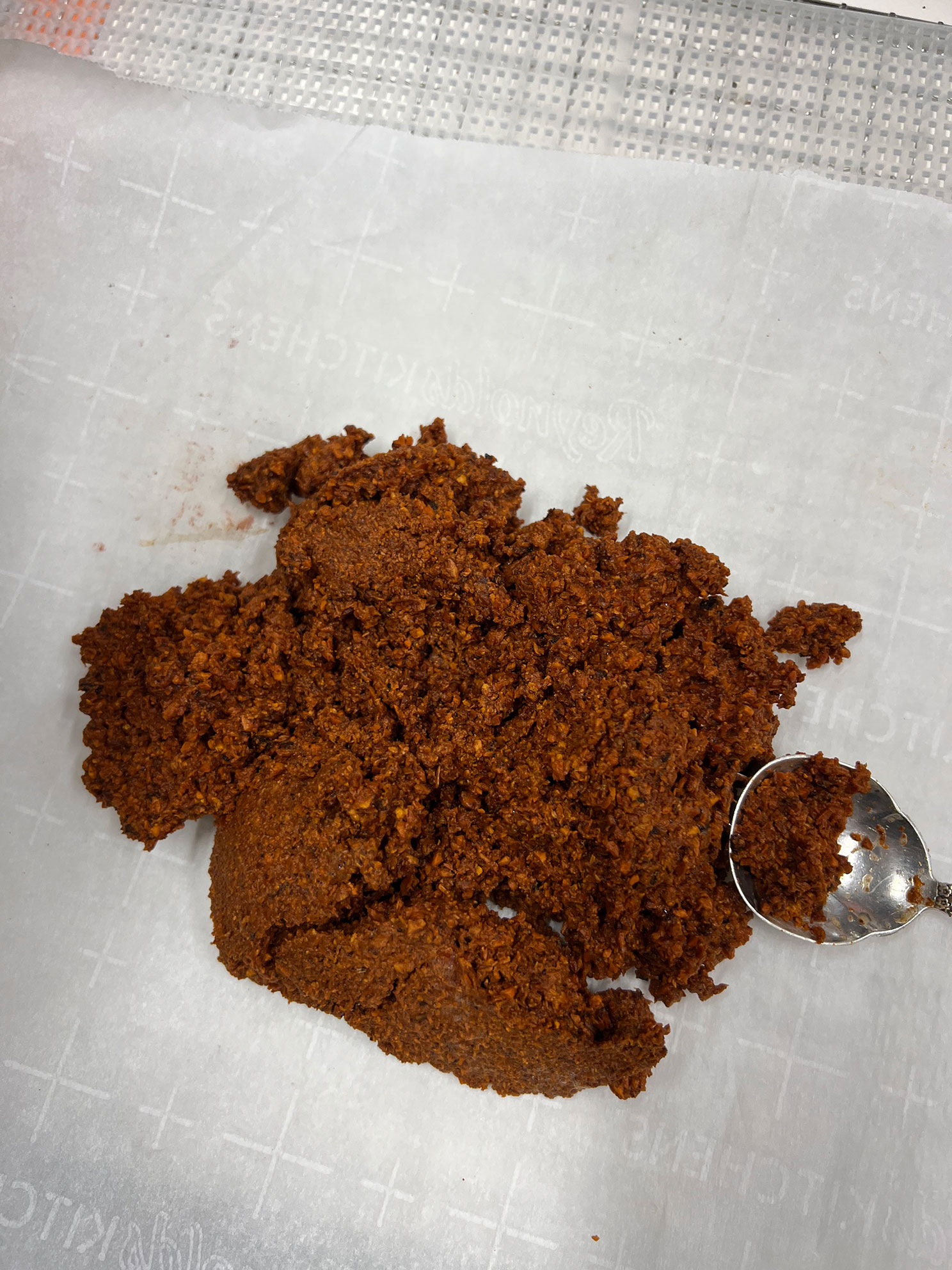 Spoon scooping brown textured paste from a pile on white parchment paper with grid lines.
