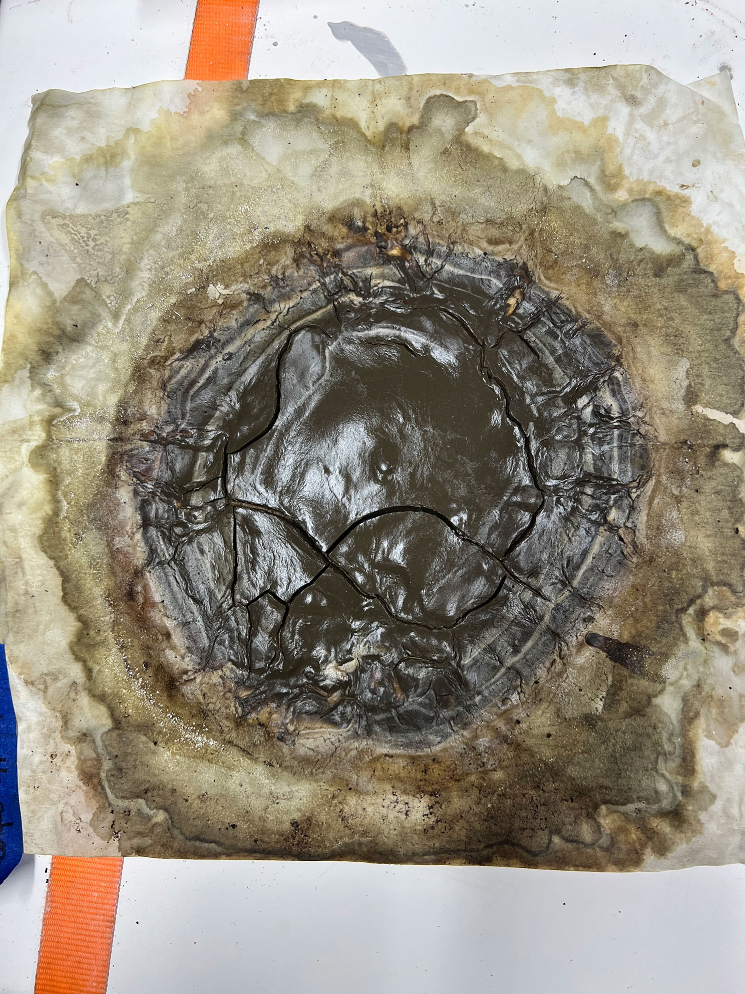 Cracked dark substance in circular pattern on stained, wrinkled parchment or paper surface.