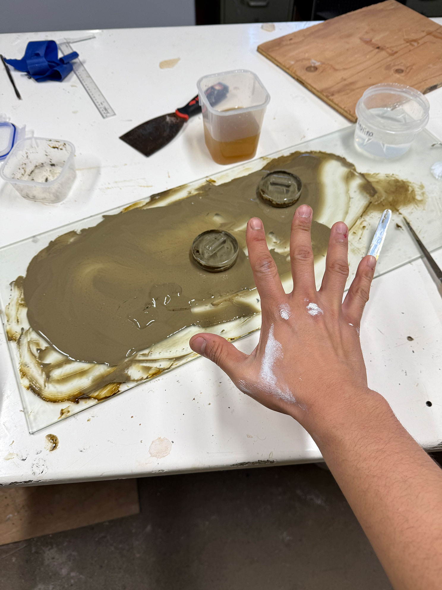 Hand with white powder marks over a glass sheet smeared with brown clay on a workbench.