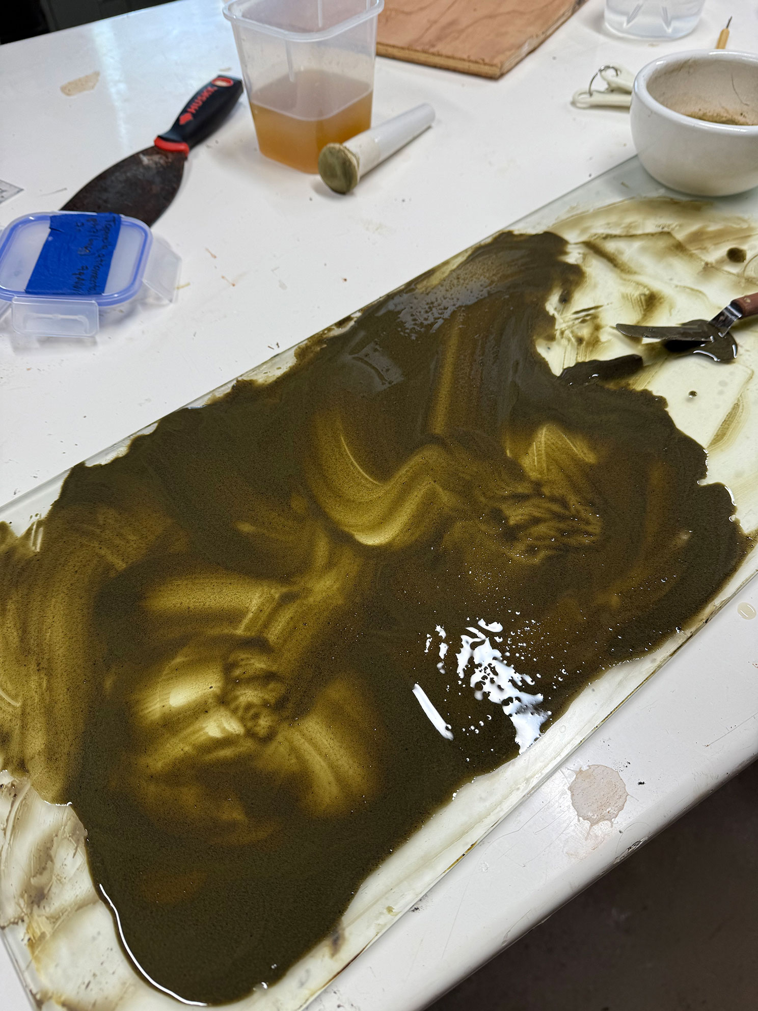 A messy work surface with dark oily or muddy liquid spread over a glass panel and nearby tools and containers.
