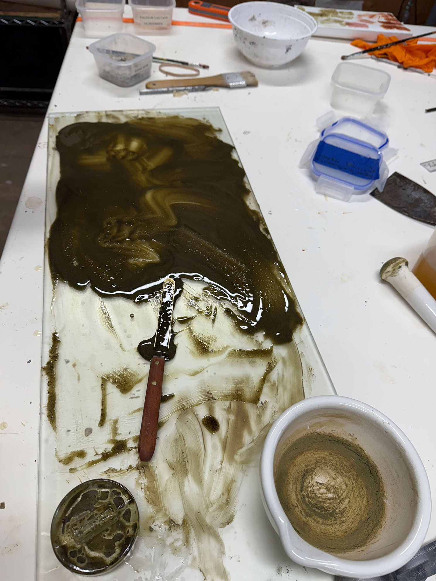 Worktable with glass plate covered in dark slime, a spatula, small containers, brush, and ceramic mortar.