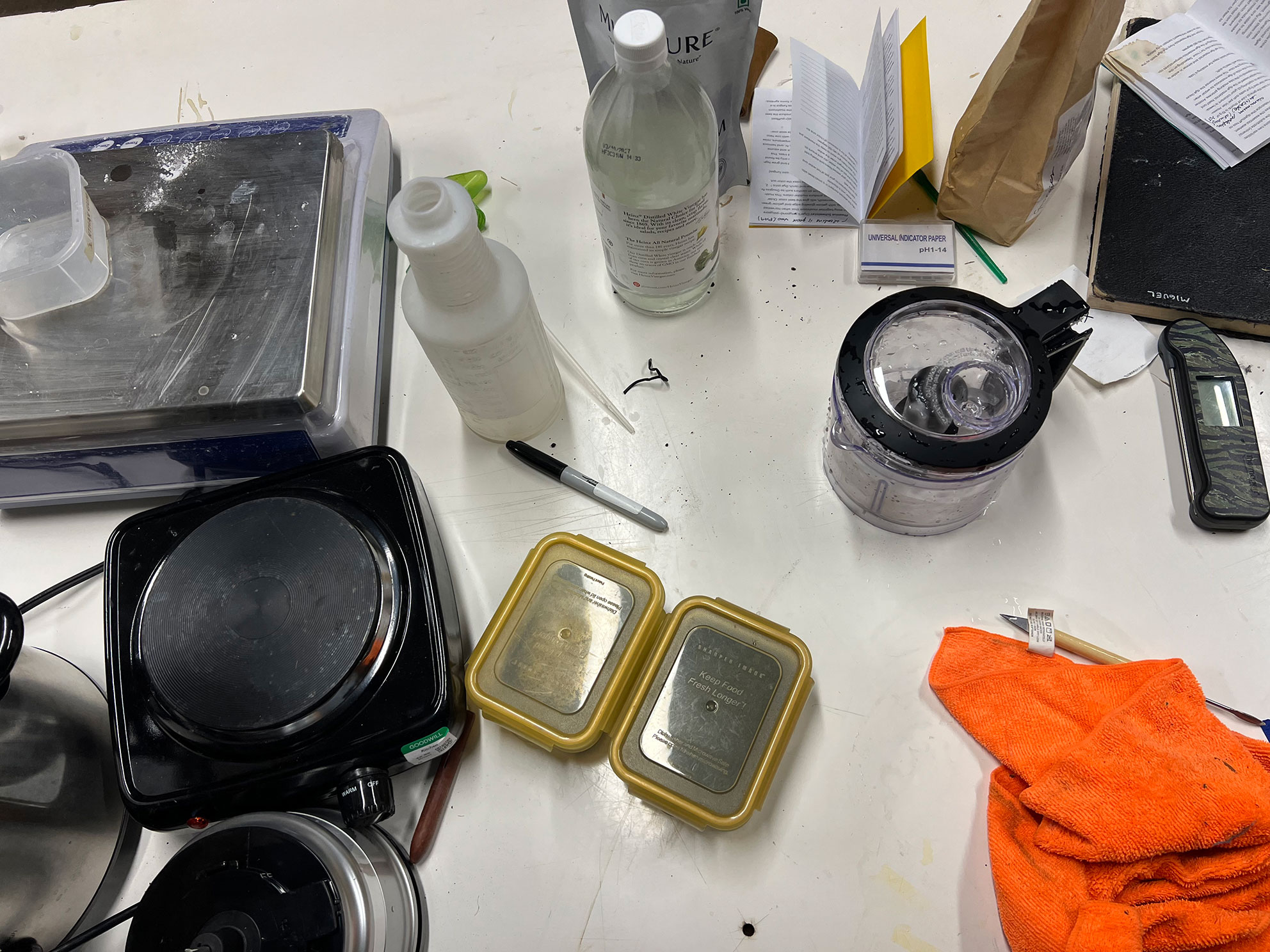 Cluttered white table with kitchen scales, containers, spray bottle, blender, pen, and orange cloth.