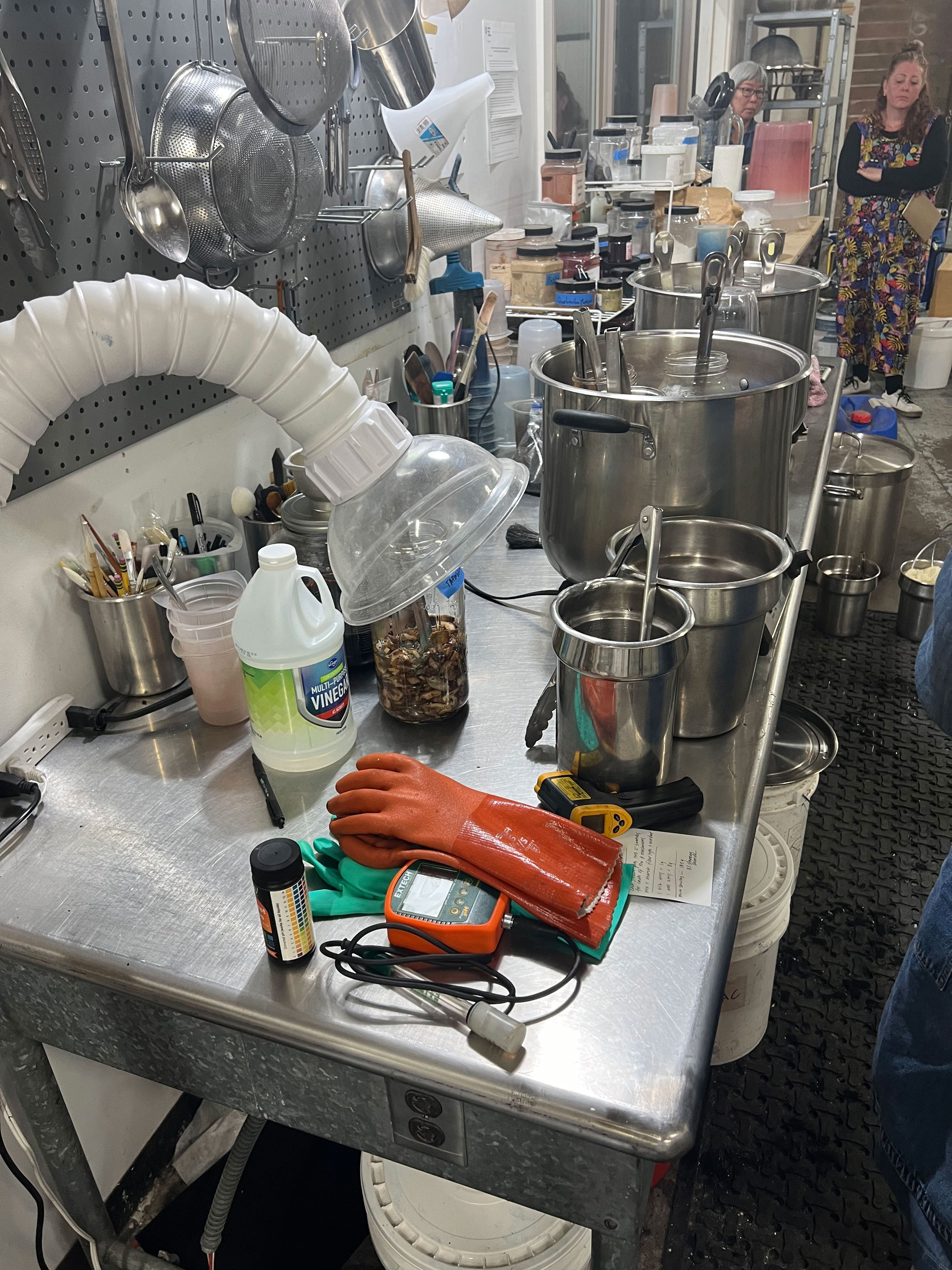 Metal table with kitchen tools, gloves, vinegar, pots, utensils, and people in background in kitchen setting.