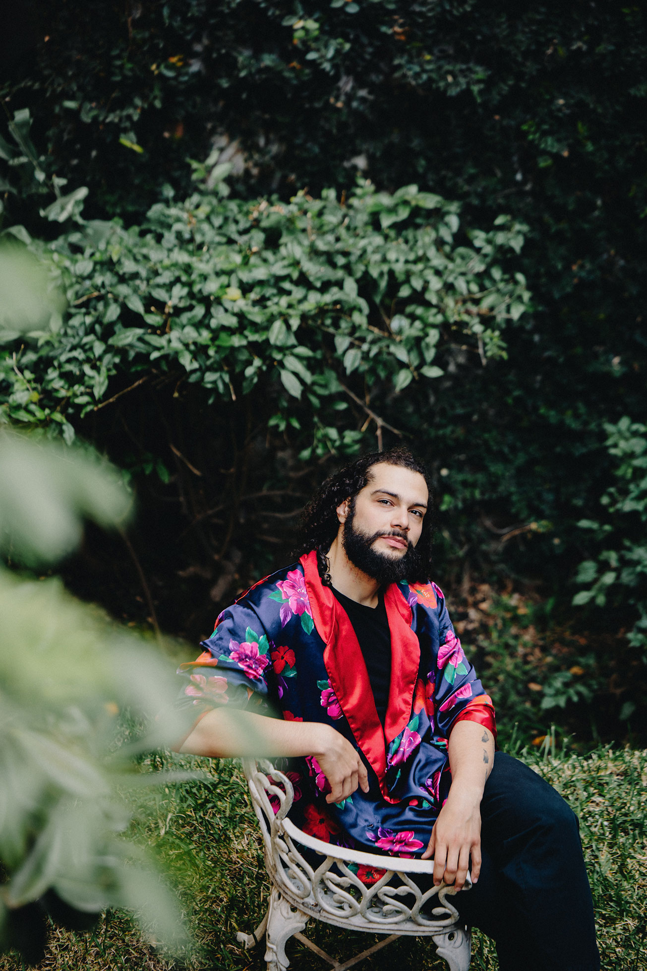 Bearded man in a floral satin jacket sitting on a white metal chair outdoors with greenery behind.